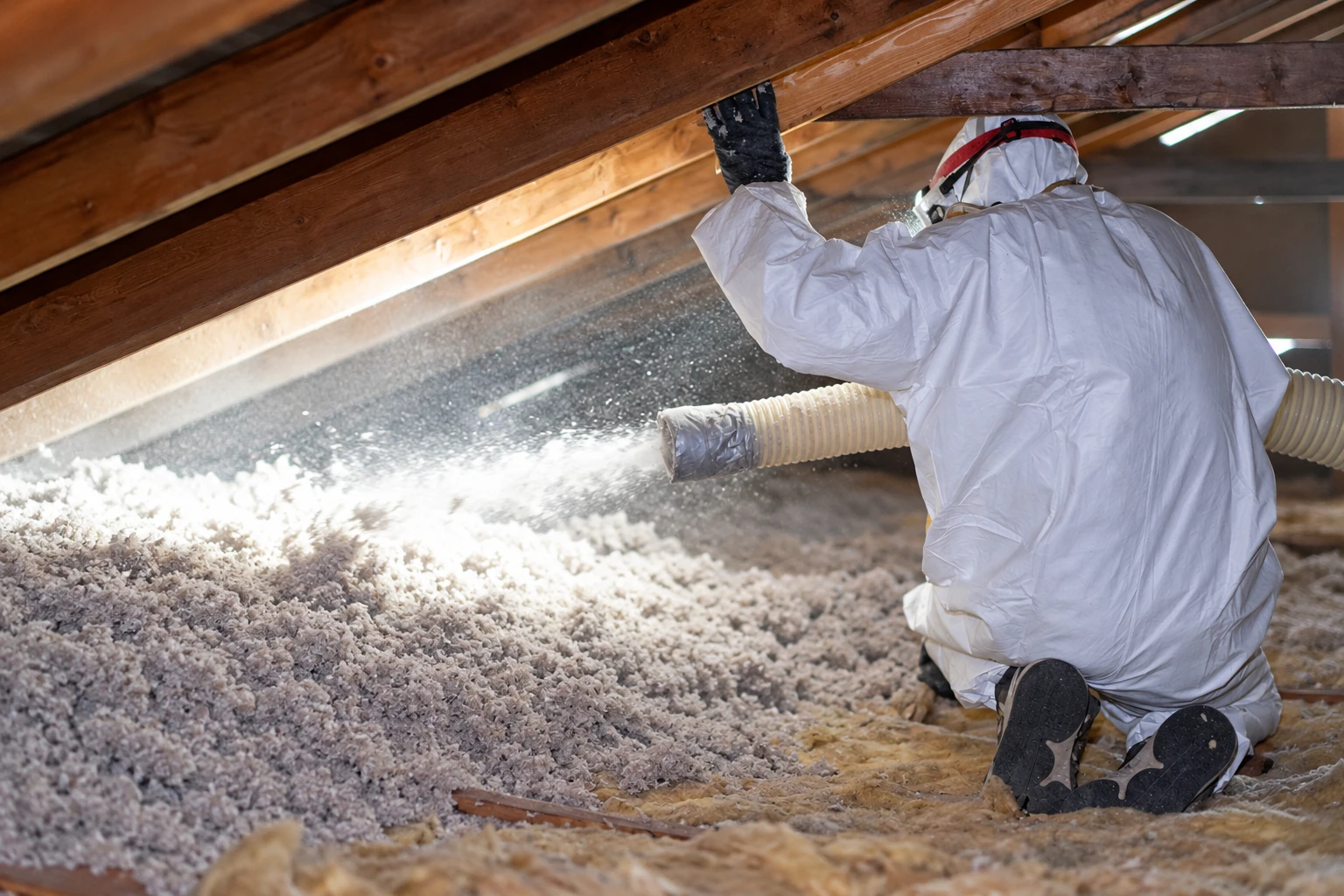 A worker in protective coveralls blows loose-fill insulation across an attic floor through a hose.