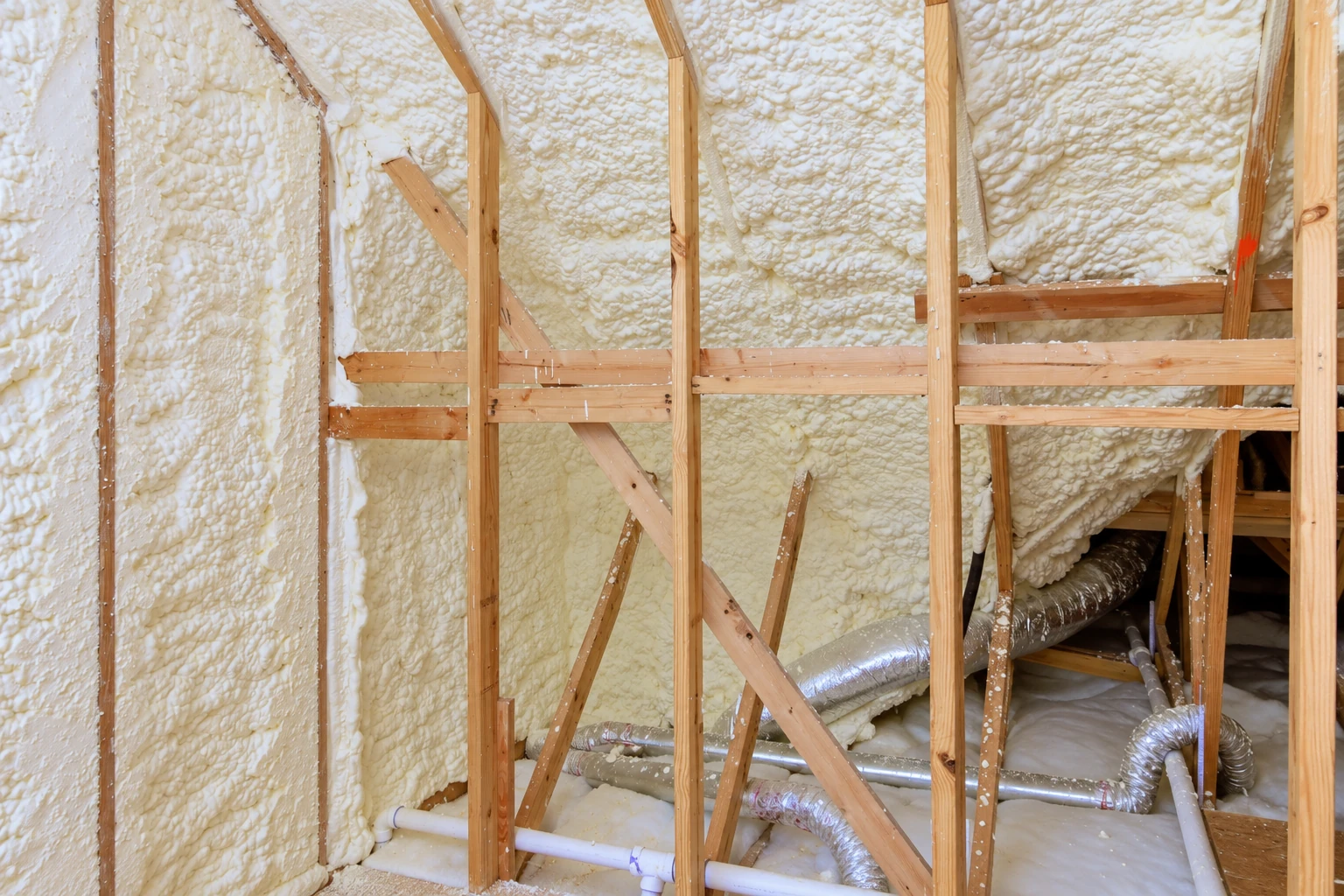 Exposed roof framing coated with thick white spray foam, with metal ducts and pipes below.