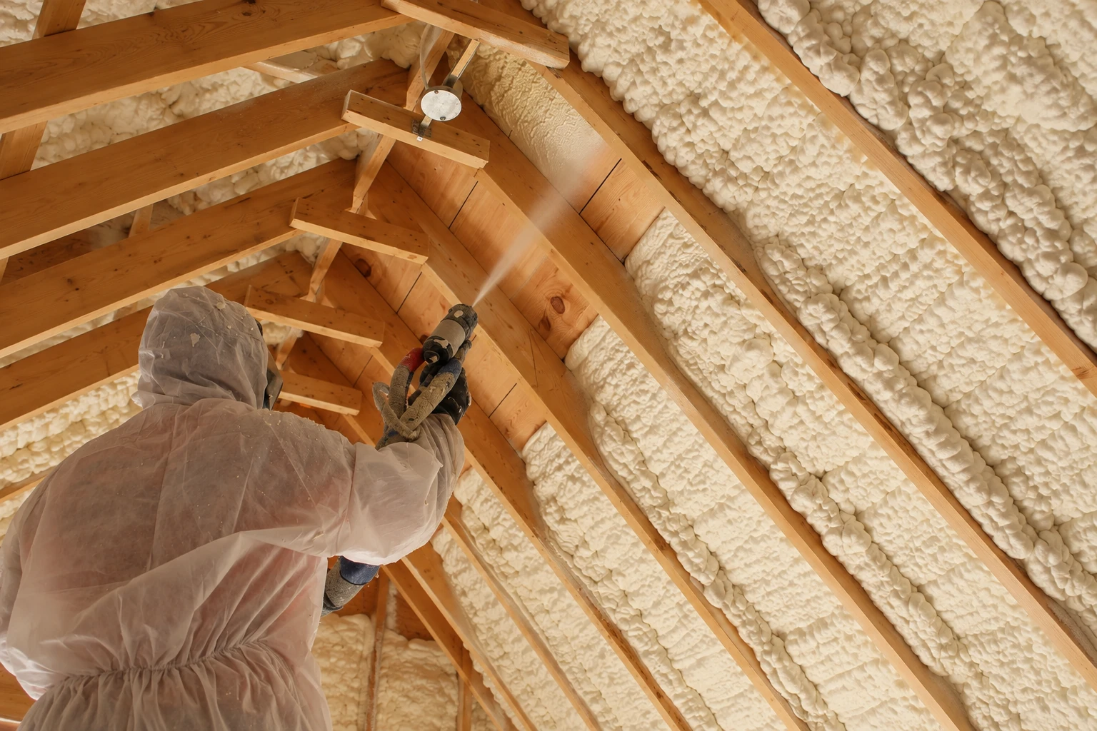 A worker in protective coveralls sprays foam insulation between the exposed rafters of a wooden roof.