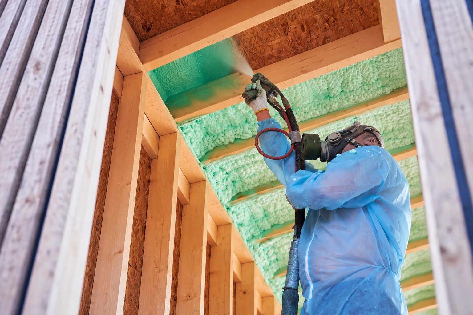 A worker in protective gear sprays green foam insulation between exposed wooden wall studs.